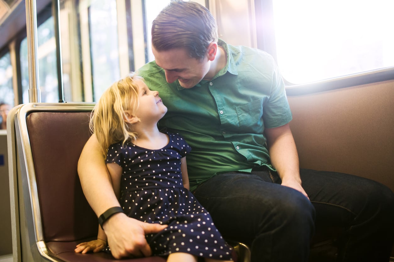 A man in a green shirt and a young girl in a polka dot dress smile and look at each other while sitting together on a train, enjoying the excitement of their latest vacation swap adventure.