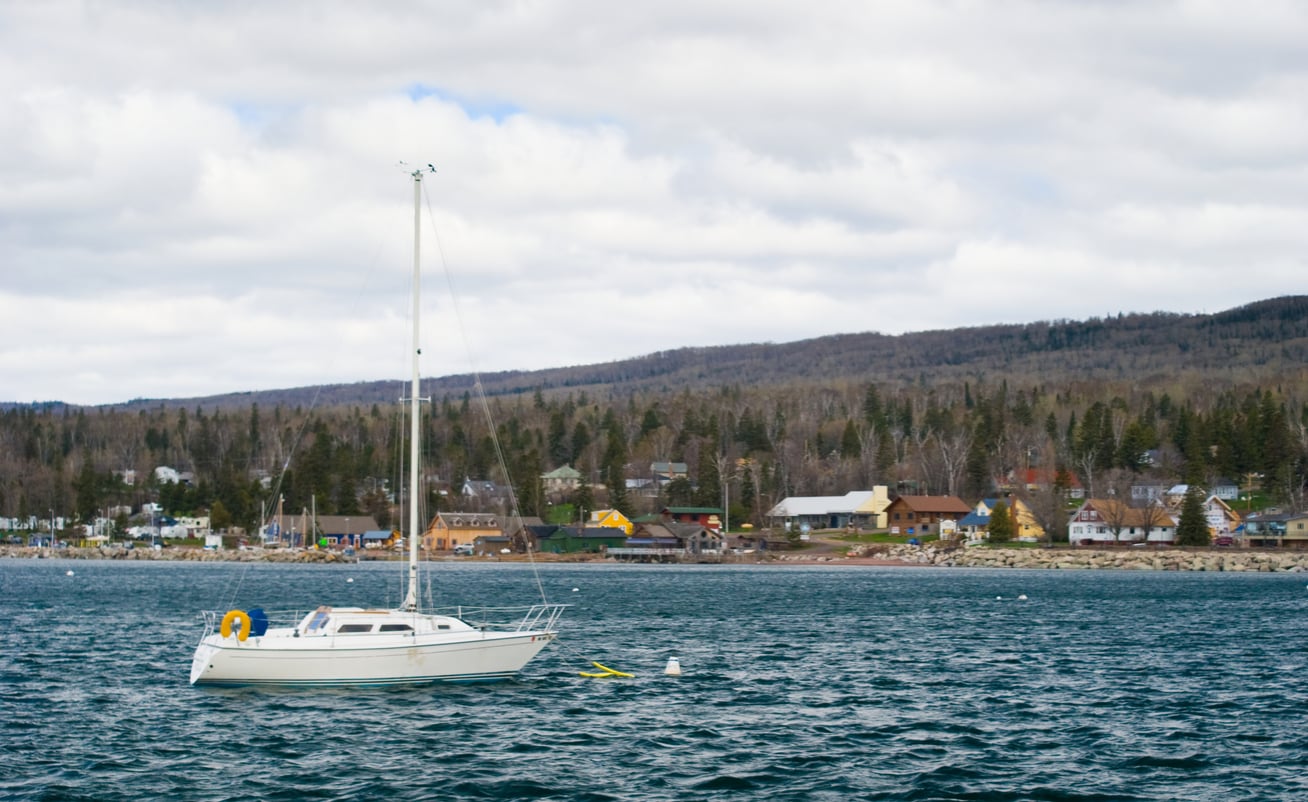 A white sailboat floats on calm blue water near a small lakeside town, with houses and trees along the shore and forested hills in the background under a cloudy sky.