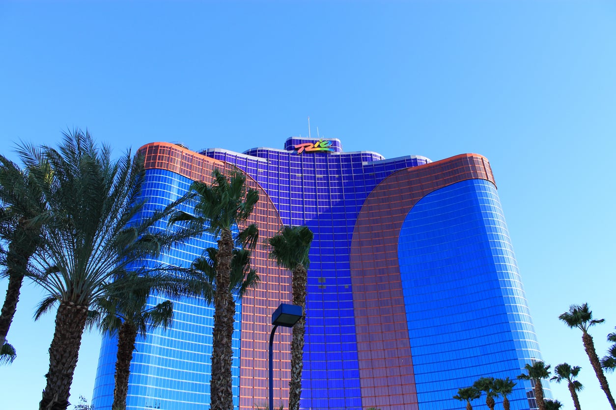 Tall palm trees in front of a large, curved building with blue and red reflective glass under a clear blue sky. The building features a colorful sign on the top.