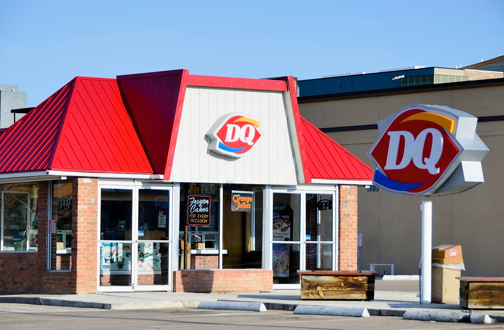 A Dairy Queen (DQ) restaurant with a red roof, brick and white exterior, and a large DQ sign out front. The windows display posters and interior lights are on. It's a sunny day.