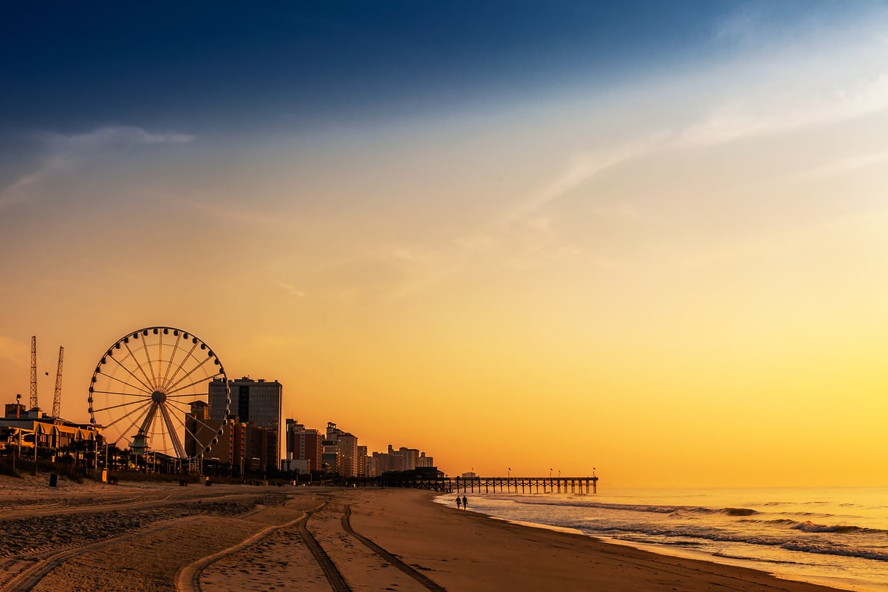 A Ferris wheel and tall buildings line a sandy beach at sunrise or sunset, with a pier extending into the ocean and a golden sky overhead.