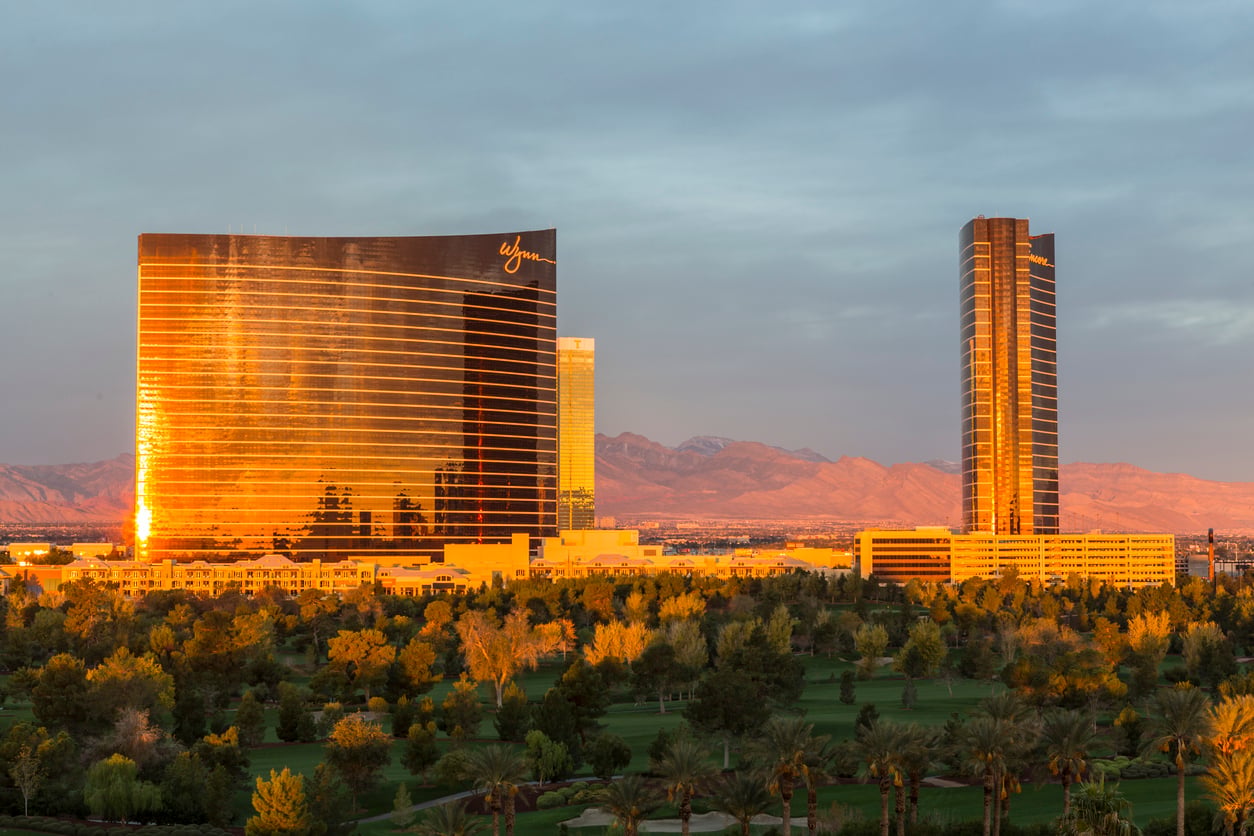 Two tall, gold-colored hotel towers reflecting sunlight, surrounded by trees and a golf course, with mountains and a cloudy sky in the background.