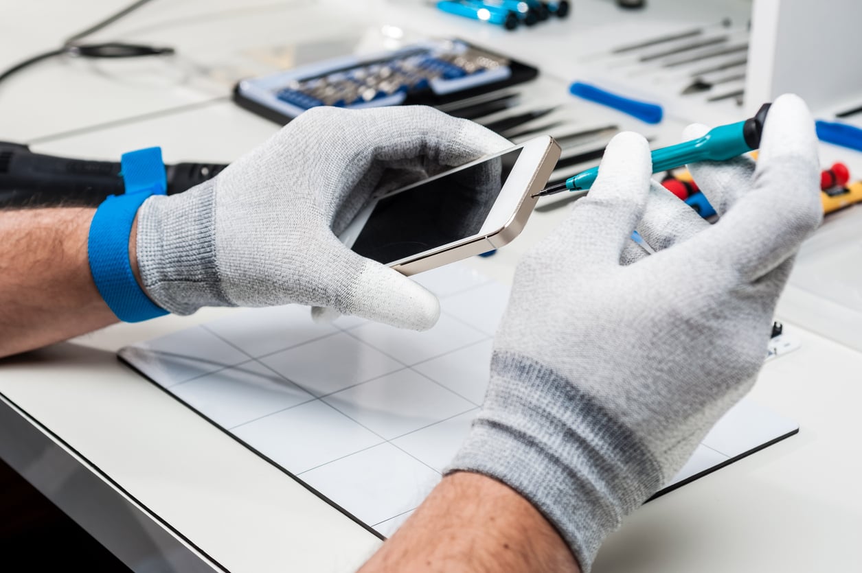 A person wearing gray gloves uses a screwdriver to repair a smartphone at a workstation with various products, tools, and parts in the background.