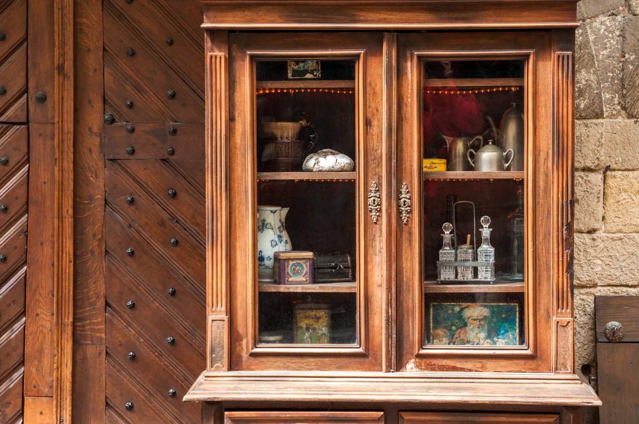 A wooden cabinet with glass doors displays vintage items, including vases, pitchers, glass bottles, a seashell, and framed pictures—evoking the nostalgic charm of Middle-Class Moms in the 80s and 90s—set against a rustic wooden and stone wall background.