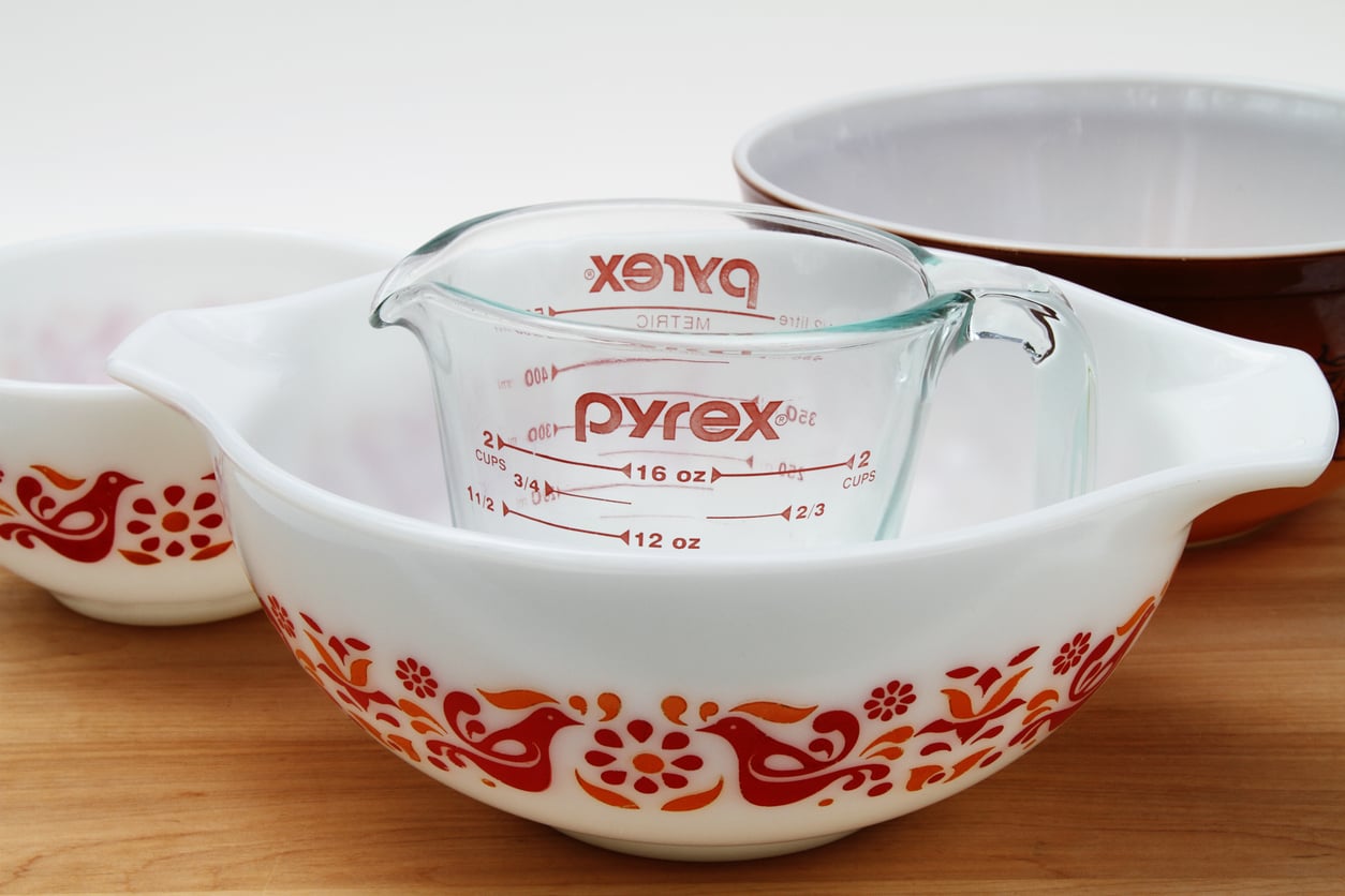A clear glass Pyrex measuring cup sits inside a white mixing bowl with a red and orange bird pattern, surrounded by other vintage kitchen items and bowls on a wooden surface.