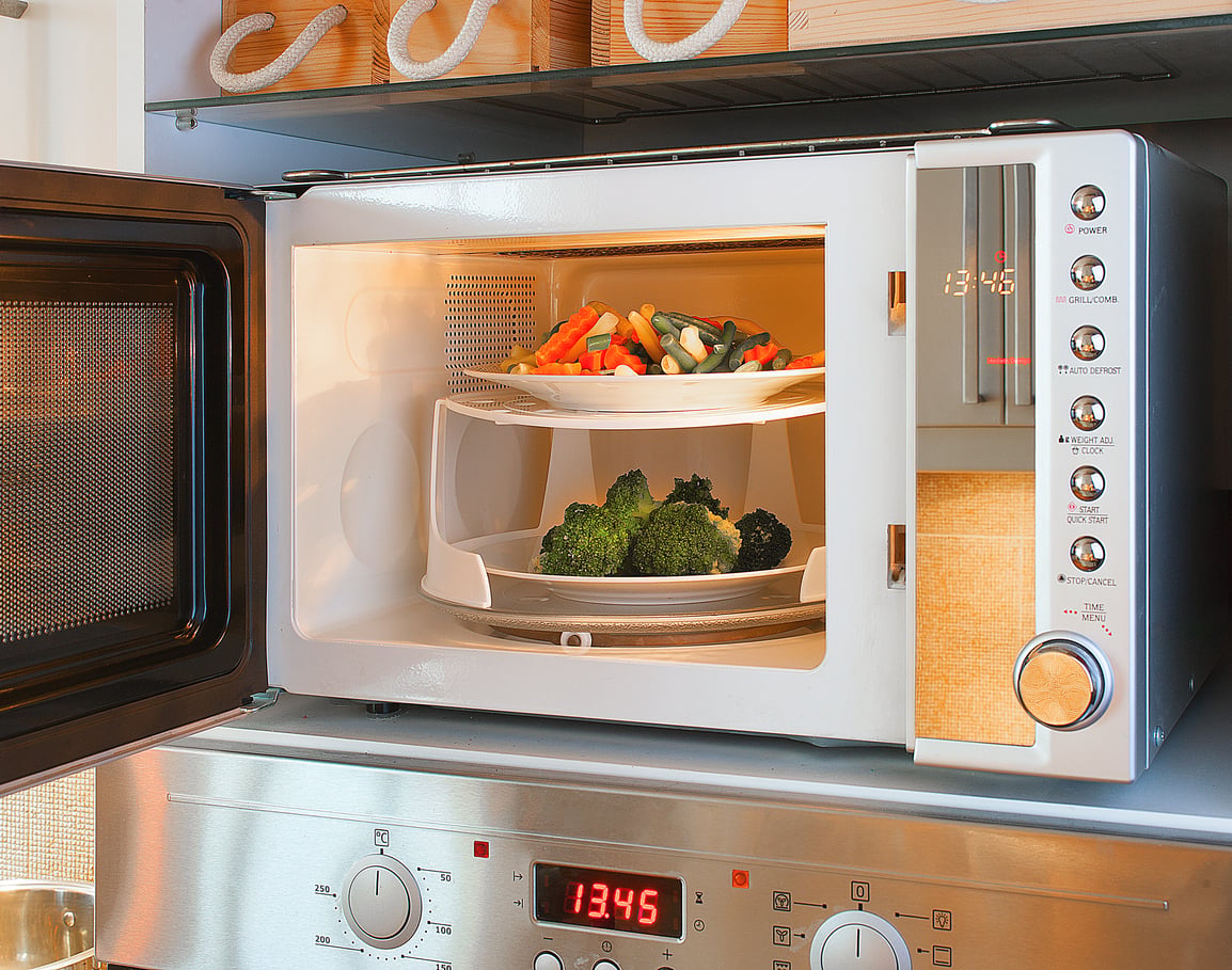 An open microwave oven with two plates of vegetables, including broccoli and mixed vegetables, inside. The microwave, one of the essential kitchen items, is placed on top of a stainless steel oven.
