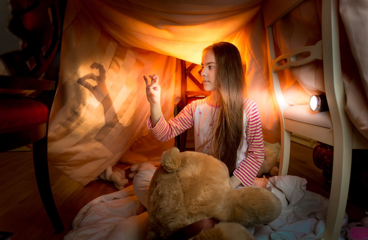 A girl sits inside a homemade blanket fort, holding a teddy bear and making shadow puppets on the wall using a flashlight. Warm light fills the cozy space, creating the perfect setting for imaginative Halloween activities.