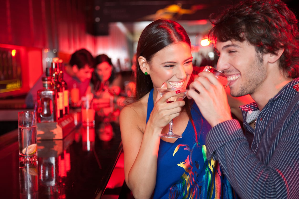 A smiling man and woman sit at a bar, enjoying drinks together. Several other people are seated in the background. The lighting is warm and red, creating a lively, festive atmosphere. Bottles are visible on the bar counter.