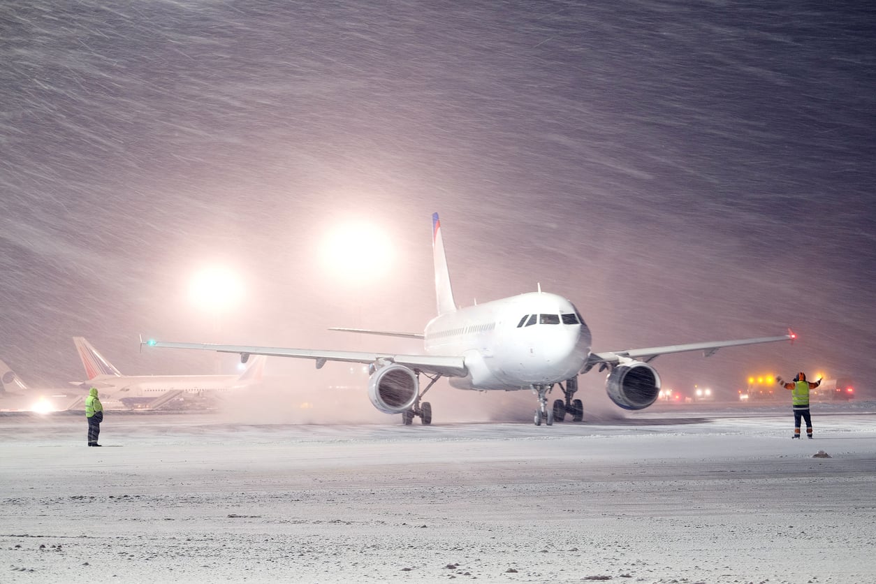 Before booking a flight, imagine an airplane on a snowy runway at night, with two ground crew members guiding it. Bright lights shine through heavy snowfall, and other planes and vehicles are visible in the background.
