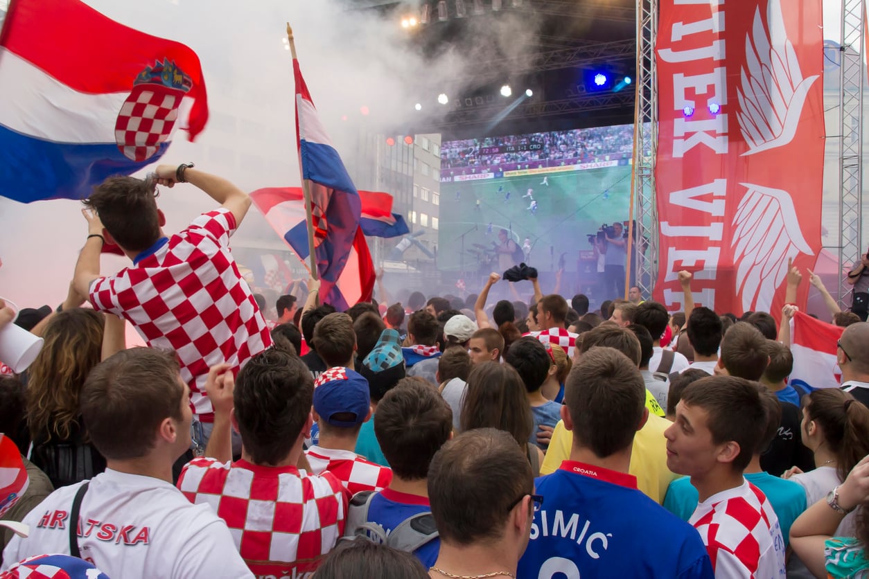 A crowd of soccer fans wearing Croatian jerseys and waving flags watch a match on a large outdoor screen, with smoke and bright lights creating a festive atmosphere.