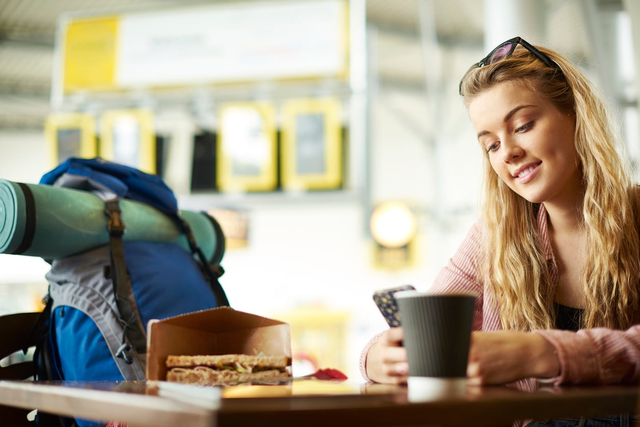 Young woman with long blonde hair smiles while looking at her phone in a cafe, perhaps checking vacation costs for her next trip. A backpack and a rolled-up mat sit beside her, ready for adventure. A sandwich and a cup are on the table in front of her. The setting is bright and modern, with yellow signage in the background.