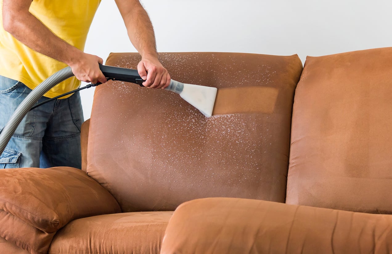 A person in a yellow shirt uses cleaning products and a vacuum machine to deep clean a brown fabric sofa, visibly removing dirt and stains from the upholstery.