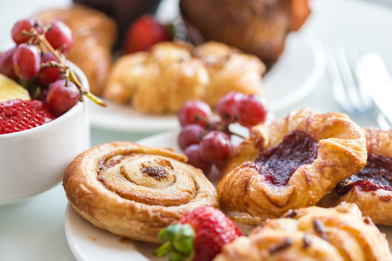 A close-up of assorted pastries, including a cinnamon roll and fruit danish, on a plate with fresh strawberries and grapes. A bowl of fruit and more pastries are blurred in the background.