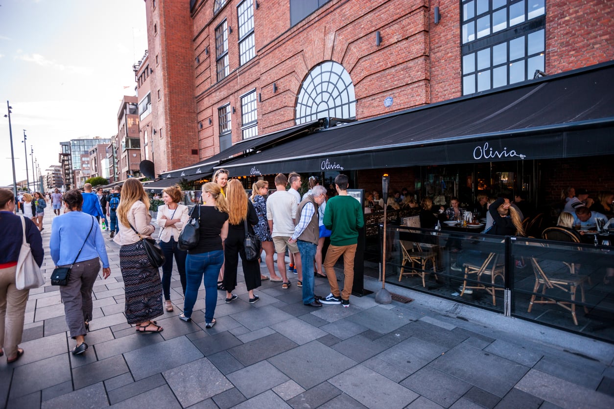 People stand in line outside a busy restaurant named “Olivia” on a city street with brick buildings, outdoor seating, and a lively atmosphere in the early evening.