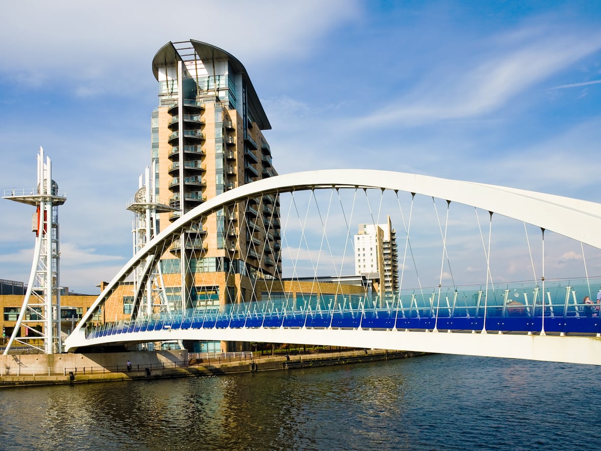Modern white arch bridge with blue accents spans a river, with a tall, glass-fronted apartment building and other contemporary structures in the background under a clear, blue sky.