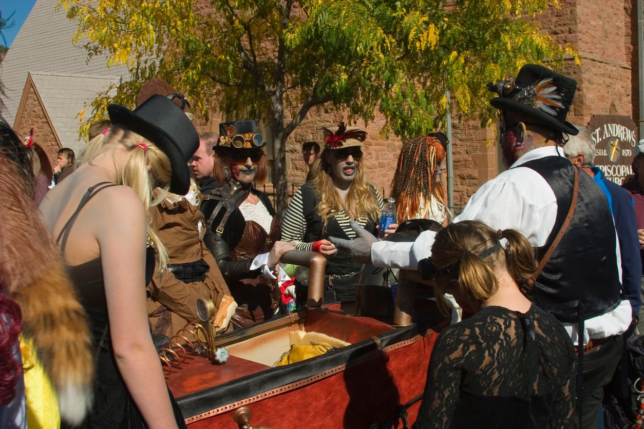 A group of people in steampunk costumes gather around a decorated cart outdoors, near a stone building, with autumn tree foliage above them, celebrating the festive spirit of Halloween.