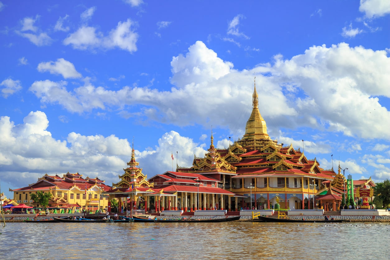 Golden pagoda with red-roofed buildings sits beside a river under a bright blue sky with scattered clouds; boats are docked along the water in front of the ornate temple complex, making it one of the region’s most captivating travel destinations.