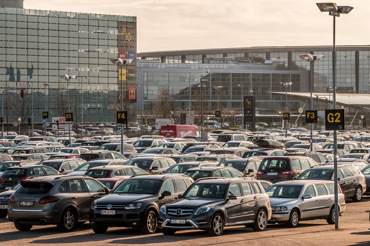 Large outdoor parking lot filled with various cars, with clearly marked section signs (G2, F4, E5). Modern glass buildings are visible in the background under a cloudy sky, hinting at a bustling area where vacation costs might be considered when choosing convenient parking options.