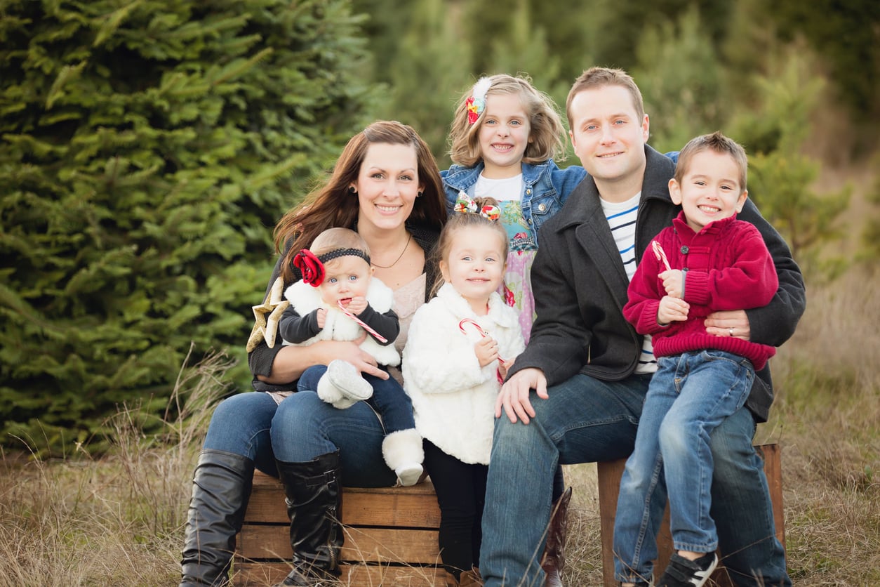 A family of six, including two adults and four young children, sits outdoors in front of pine trees. They are smiling and dressed in cozy winter clothing, enjoying quality time together without worrying about vacation costs.
