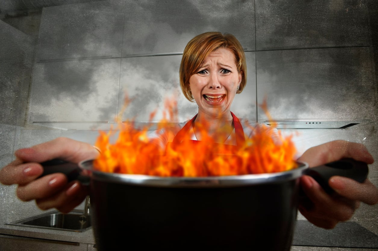 A woman in a kitchen holds a flaming pot, looking distressed and worried as smoke rises in the background.