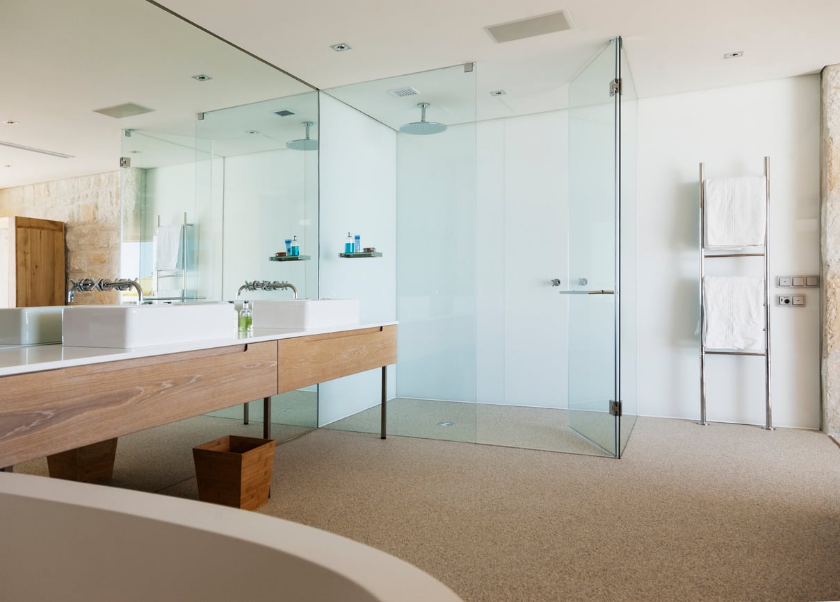 Modern bathroom with a large glass-enclosed shower, double sinks on a wooden vanity, wall-mounted towel rack with white towels, and neutral-toned flooring. The space is bright and minimalistic.