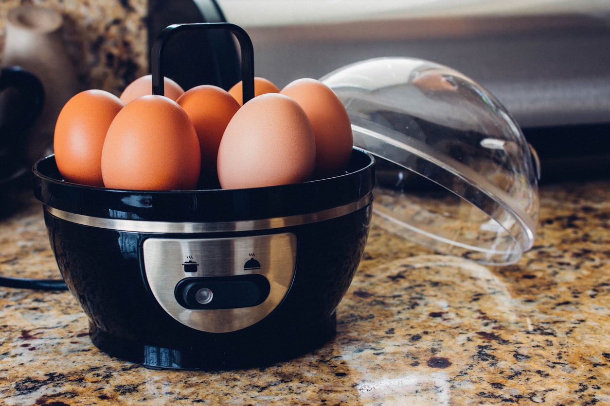 A black electric egg cooker, one of the must-have kitchen gadgets, sits on a granite countertop holding seven brown eggs. Its clear plastic lid is resting beside it, and the power switch is visible on the front of the appliance.