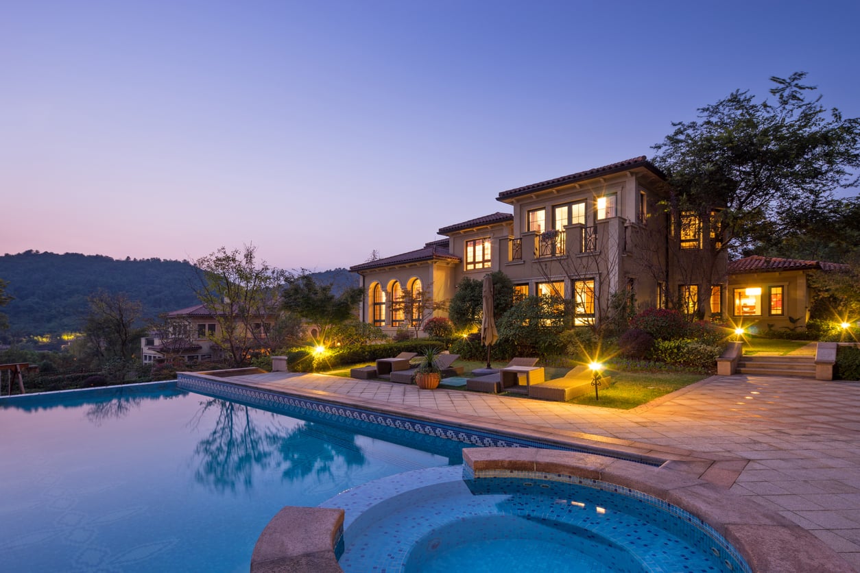 A luxurious house with large windows is lit up at dusk, overlooking a spacious swimming pool and hot tub, surrounded by outdoor seating, trees, and distant hills under a clear evening sky.