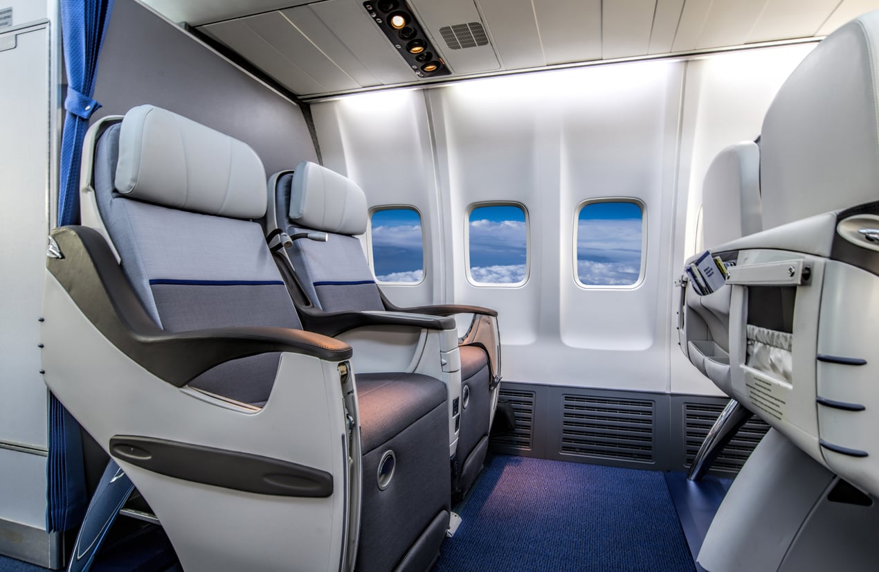 Two modern airplane seats by three windows with a view of clouds and blue sky; spacious legroom and a clean, comfortable interior.