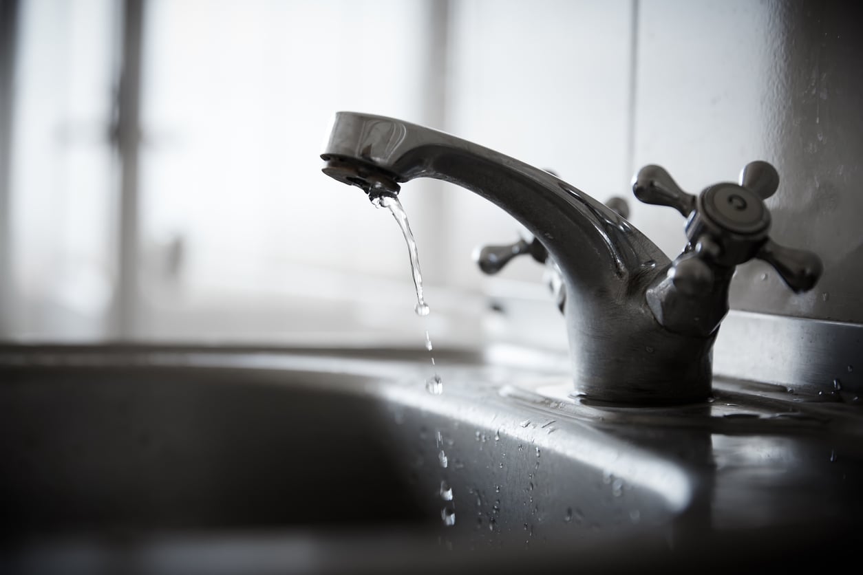 A close-up of a metal kitchen faucet with a few drops of water dripping from the spout into a stainless steel sink, with a blurred background.