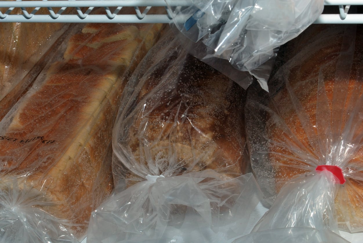 Frozen food products. Three loaves of bread wrapped in plastic bags are stored on a freezer shelf. Some frost is visible on the bags and bread, and the loaves are placed side by side.