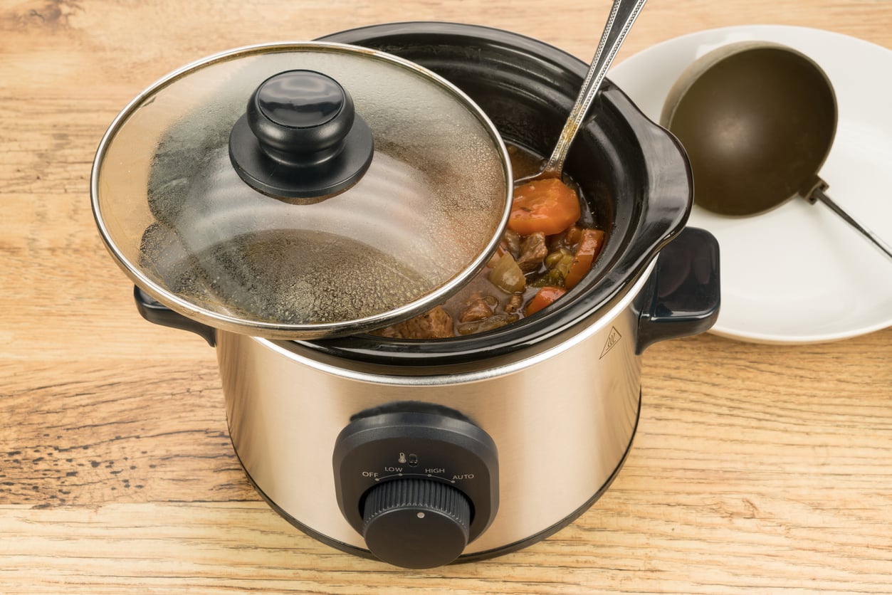 A slow cooker with its lid partly open reveals a stew with vegetables and meat; a ladle rests inside, and a white plate with another ladle, both essential kitchen items, sits nearby on a wooden surface.