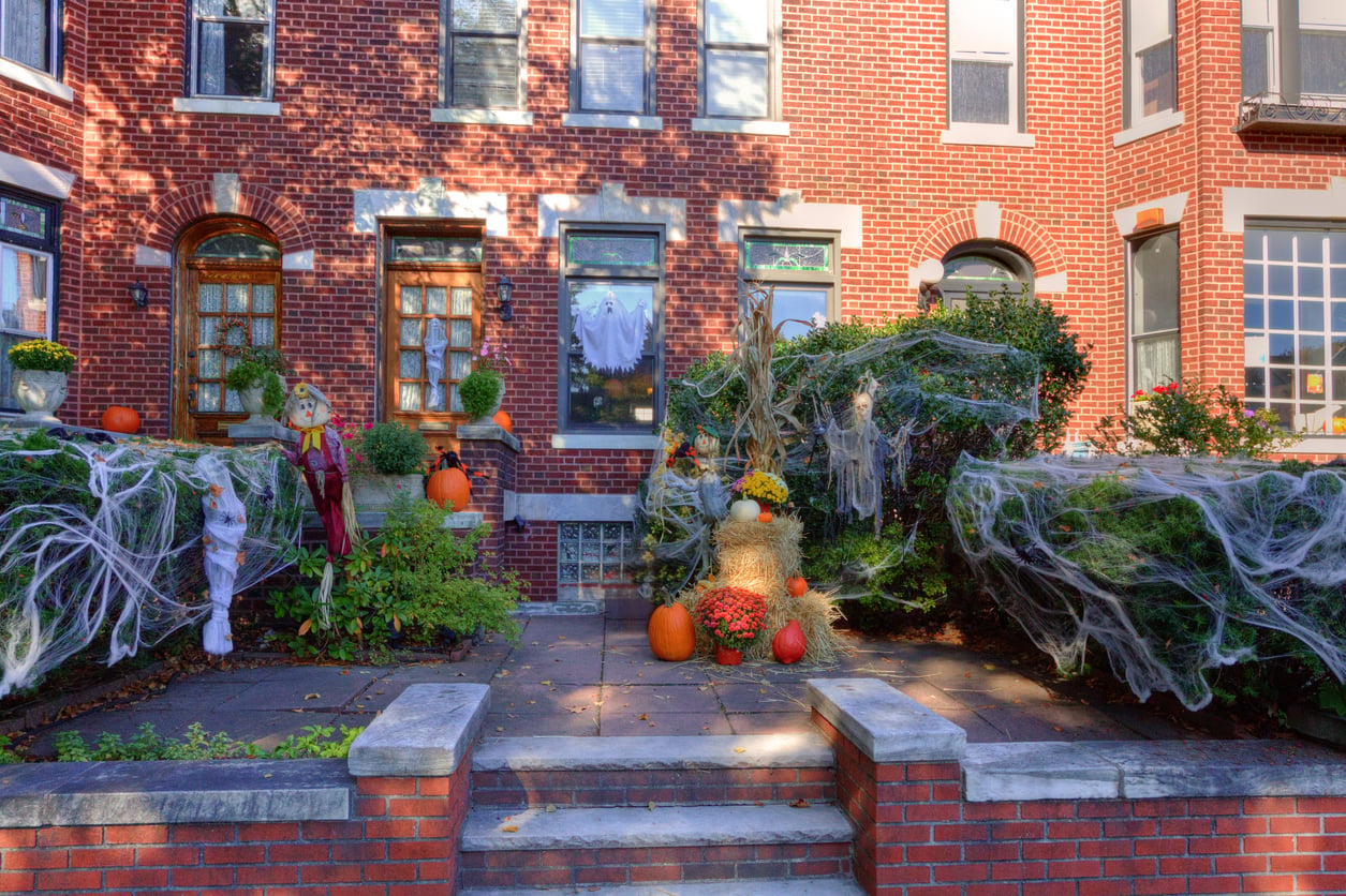 A brick townhouse decorated for Halloween in the style of the best trick or treat towns in America, with fake spider webs, pumpkins, hay bales, ghosts, and a scarecrow nestled among the bushes and plants near the entrance steps.