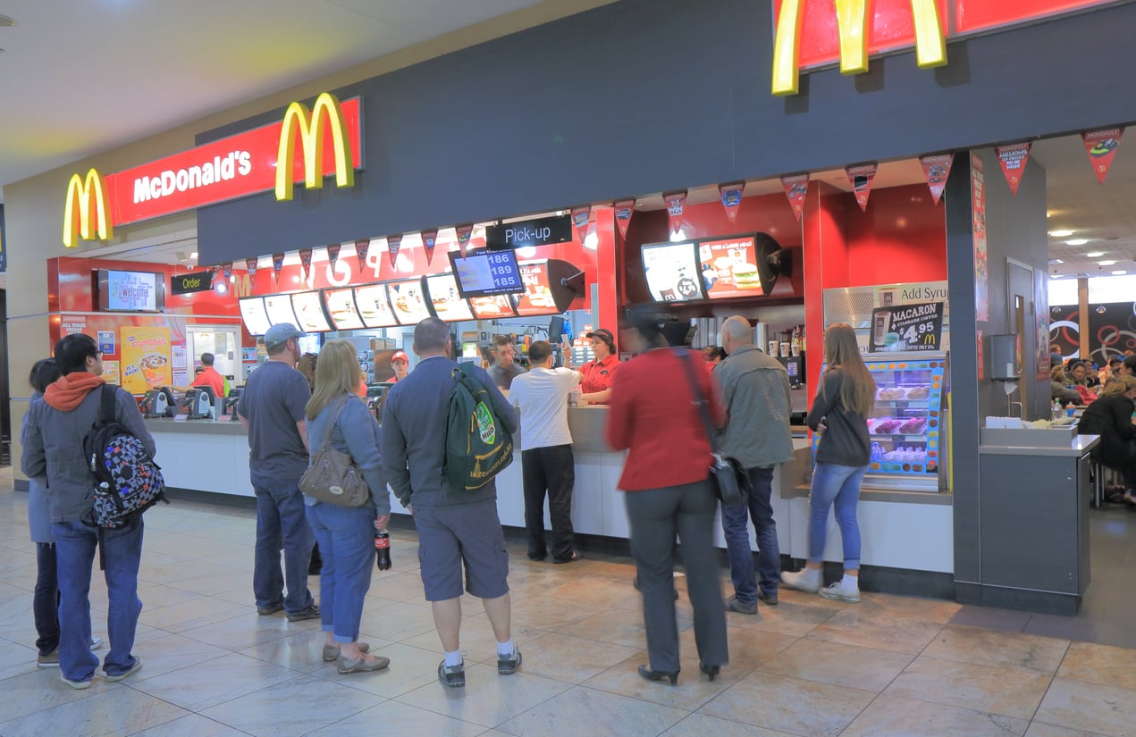 Customers stand in line at a McDonald's counter inside a shopping mall, waiting to order food. Bright menu boards display meal options above the counter, and a pickup area is visible.