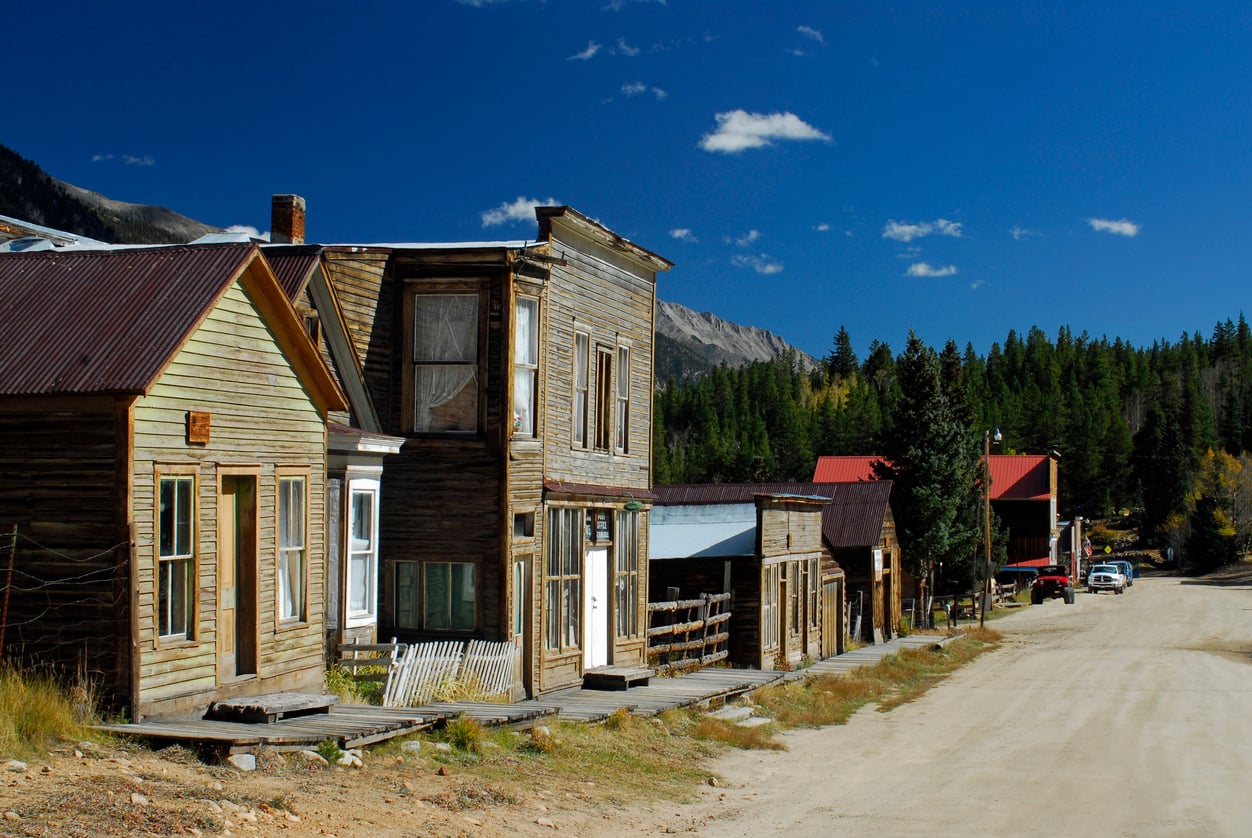 A row of old wooden buildings lines a dirt road in a small, rustic American town surrounded by trees and mountains under a clear blue sky.