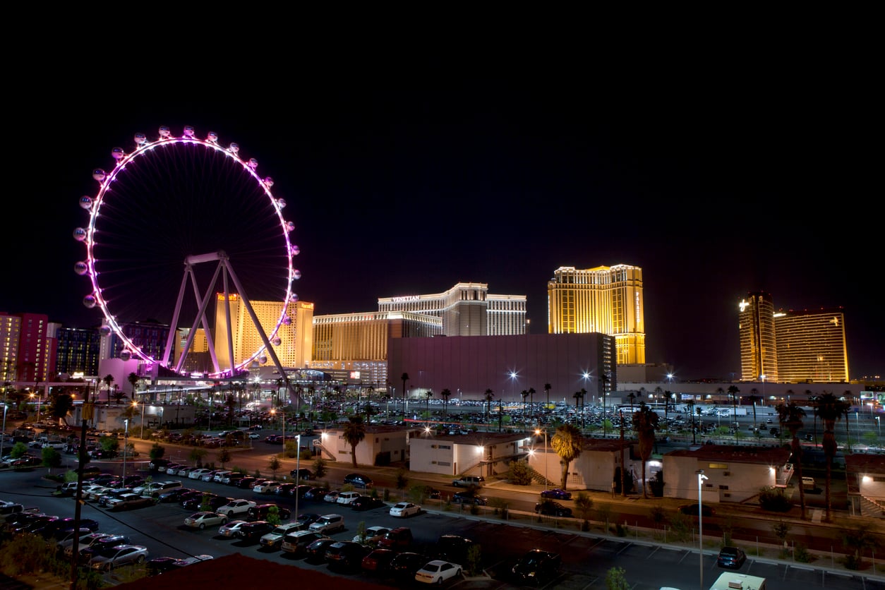 Las Vegas skyline at night featuring the illuminated High Roller Ferris wheel, brightly lit hotels, and a parking lot with numerous cars in the foreground.