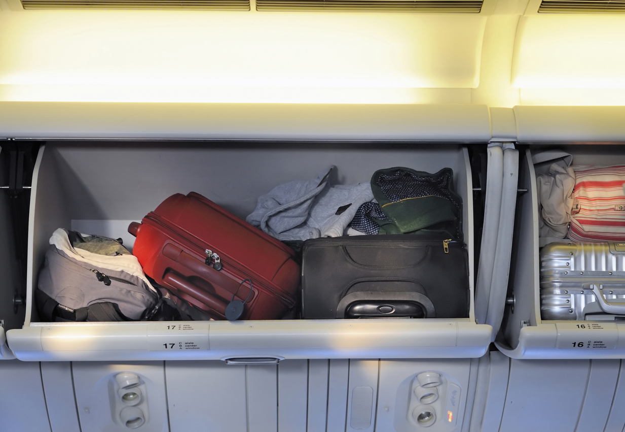 Several pieces of luggage, including a red suitcase, a black bag, and clothing, are stored in an overhead compartment on an airplane. The compartment doors are open, and the bags are tightly packed.