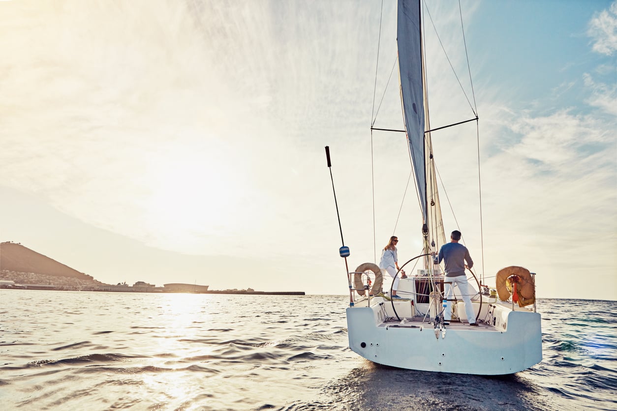 Two people stand on the deck of a white sailboat on calm water at sunset, enjoying one of life’s most expensive hobbies as the sun hangs low in the sky and a coastline is visible in the distance.