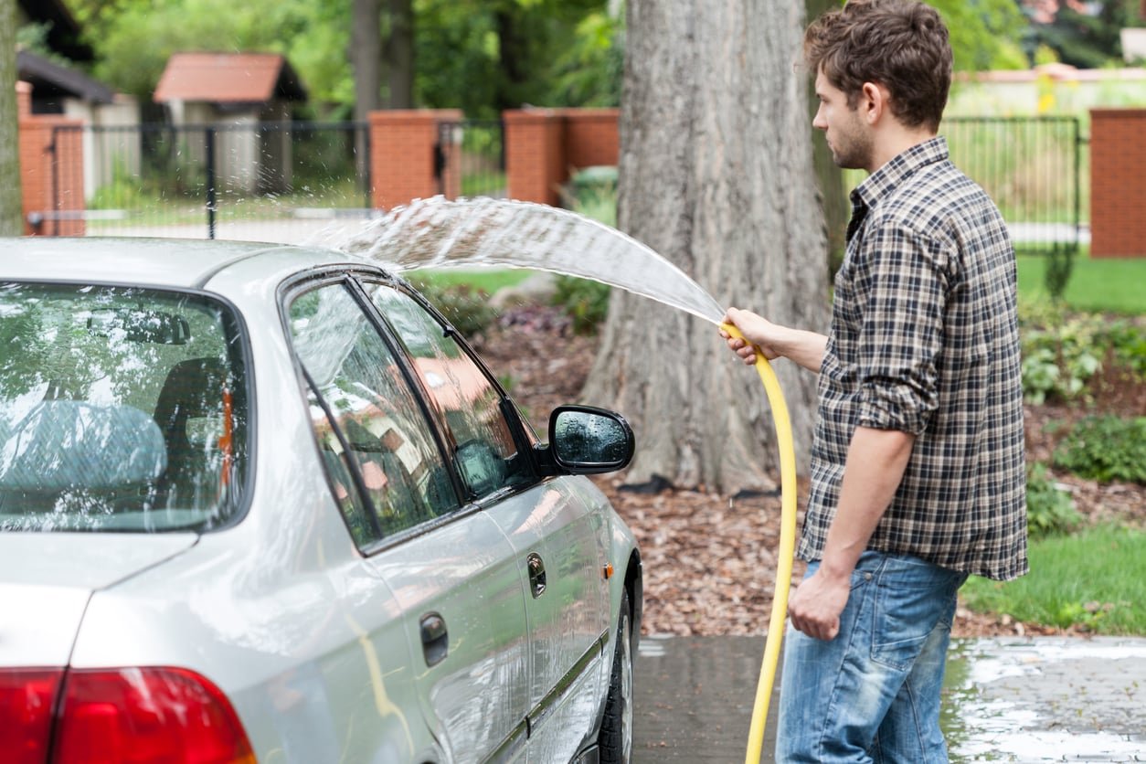 A man in a plaid shirt uses a yellow hose to wash a silver car outdoors in a leafy residential area, with trees and a brick fence in the background.
