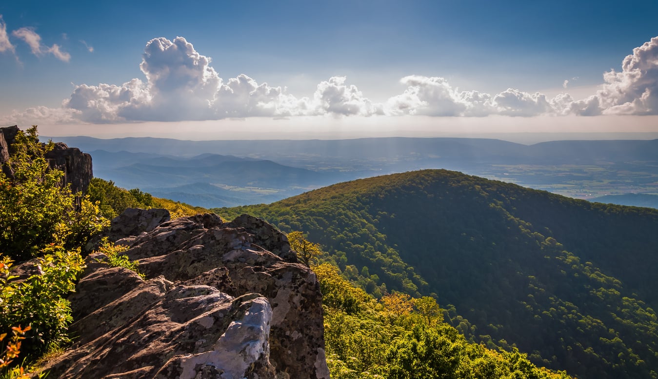 A rocky outcrop overlooks a lush, green mountain range on the East Coast under a blue sky with scattered clouds and sun rays streaming through.