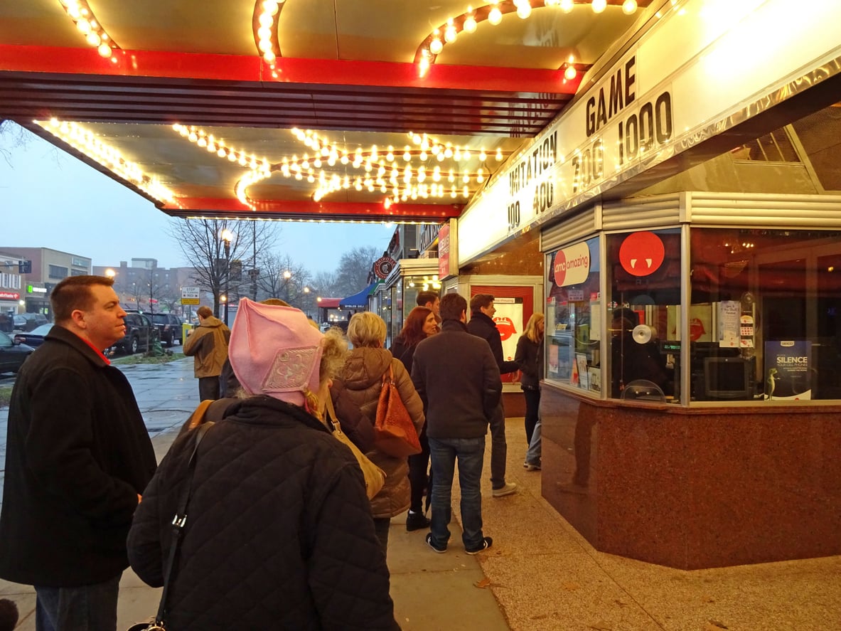 People stand in line outside a theater under a brightly lit marquee, waiting to buy tickets at the box office on a rainy evening. The sidewalk glistens, and seasoned moviegoers share movie theater hacks while buildings loom in the background.