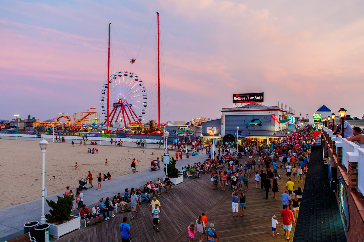 A busy boardwalk by the beach at sunset, with crowds of people, amusement rides including a Ferris wheel, bright lights, and a Ripley's Believe It or Not! museum.
