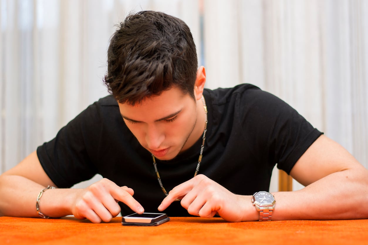A young man in a black shirt leans over a table, focusing intently as he uses both hands to operate a smartphone placed in front of him.