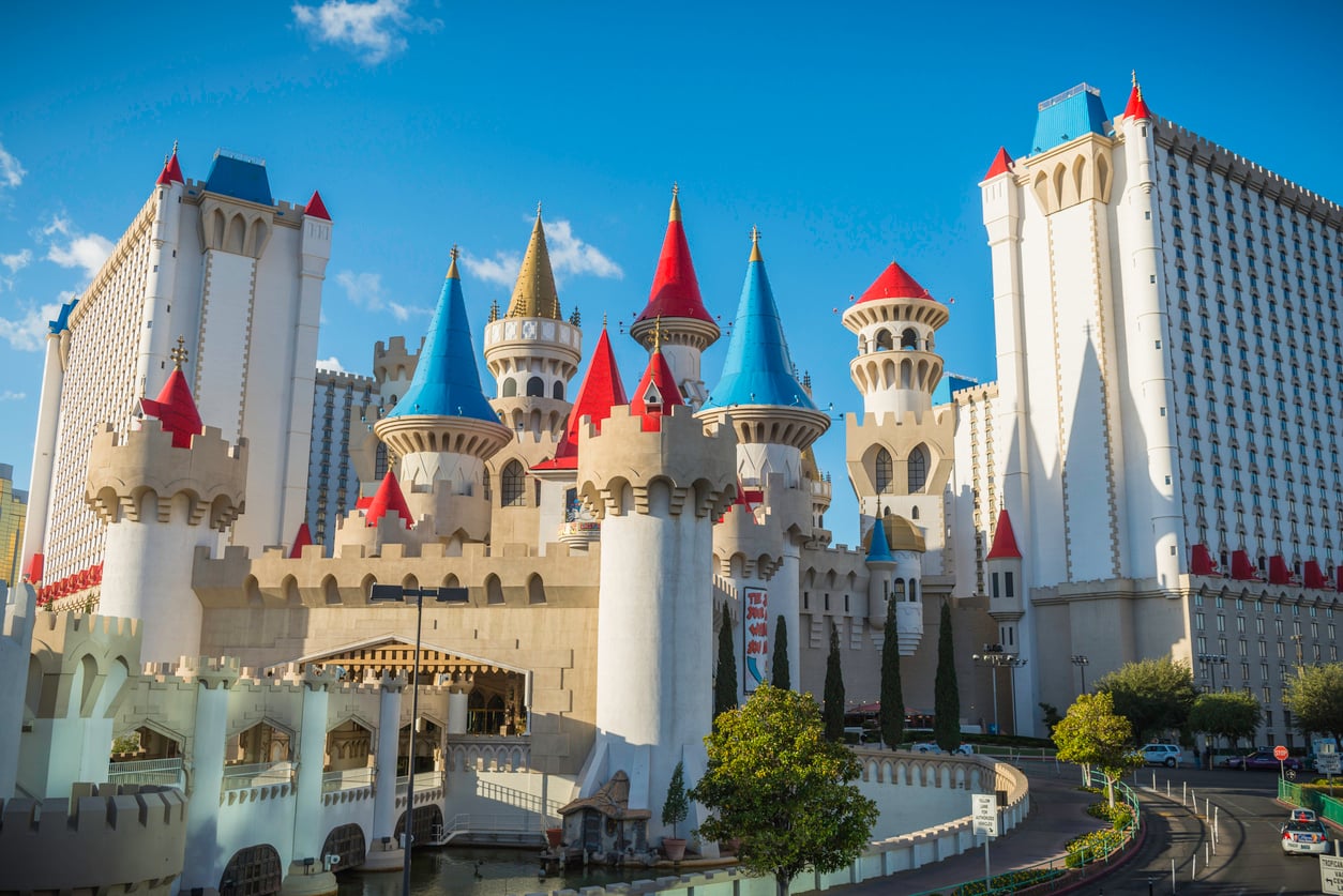 Colorful castle-themed hotel with tall blue, red, and gold turrets, surrounded by modern white high-rise buildings under a bright blue sky, with trees and cars in the foreground.