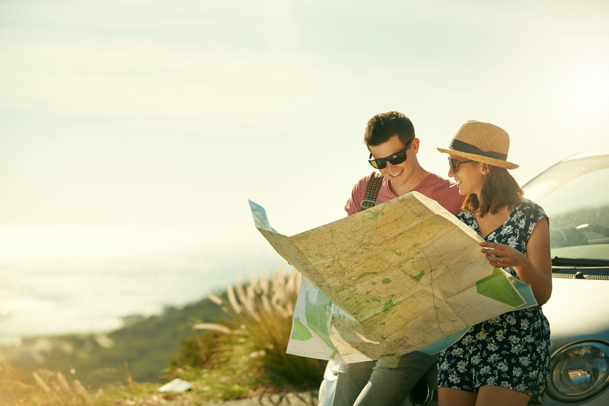 A smiling young couple leans on a car, looking at a large map together under bright sunlight, capturing the spirit of life before smartphones, with nature and blurred greenery in the background.