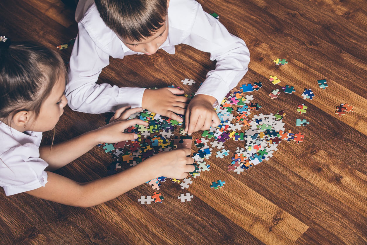 Two children in white shirts are lying on a wooden floor, working together to assemble a colorful jigsaw puzzle. The puzzle pieces are scattered between them as they focus on finding pieces that fit.