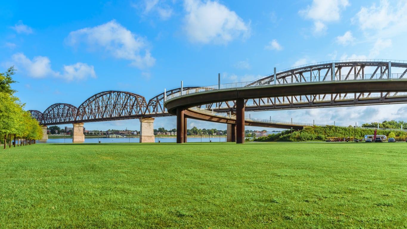 A large iron bridge with arched trusses crosses a river, while a modern curved ramp leads up to it. The scene features a wide, green grassy field under a blue sky with scattered clouds.