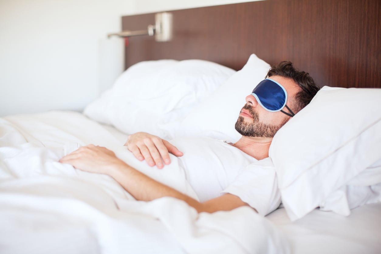 A man wearing a blue sleep mask lies on his back in bed, covered with white sheets and pillows, resting peacefully.