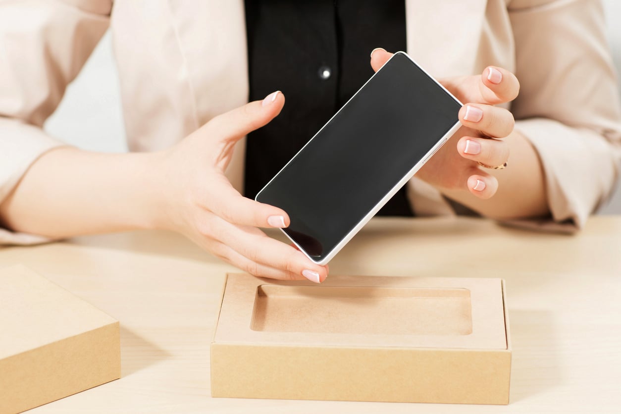 A person is holding a new smartphone above an open cardboard box on a light-colored table, preparing to take it out—highlighting how products people no longer buy, like traditional flip phones, have given way to the latest technology.