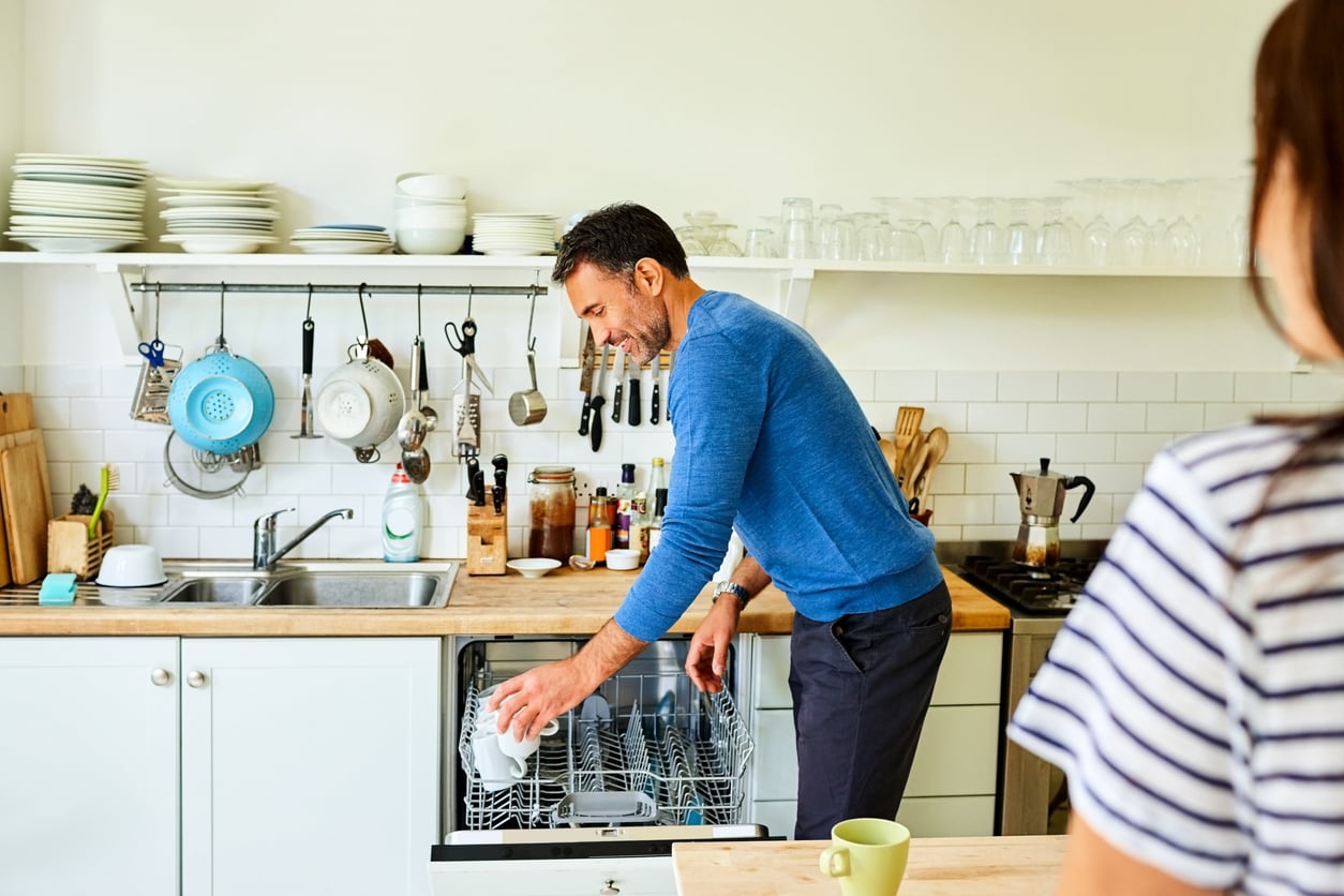 A man in a blue sweater loads a dishwasher in a bright kitchen with white cabinets, wooden countertops, and shelves holding plates, glasses, and utensils. Another person stands in the foreground, partially visible.