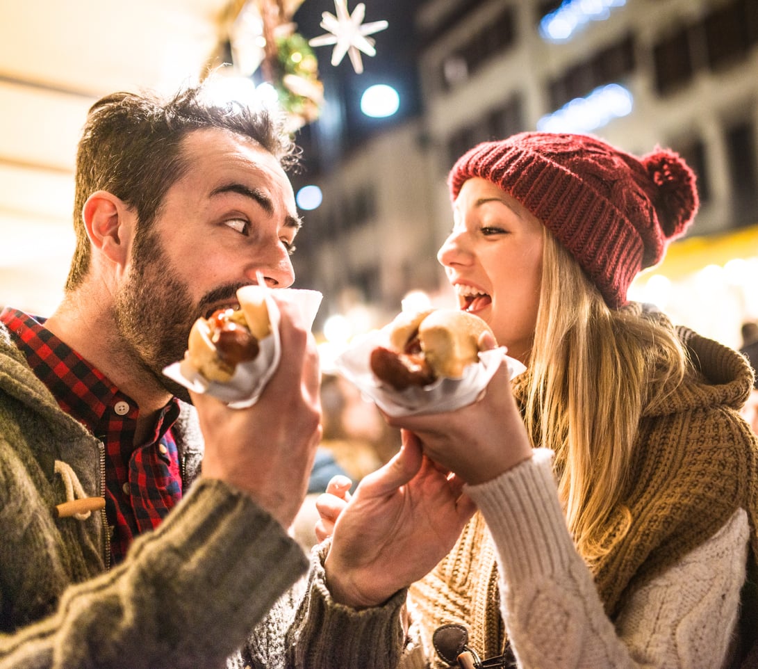 A man and woman smile and hold up sausages in buns at an outdoor event, wearing cozy winter clothing at night with festive lights in the background.